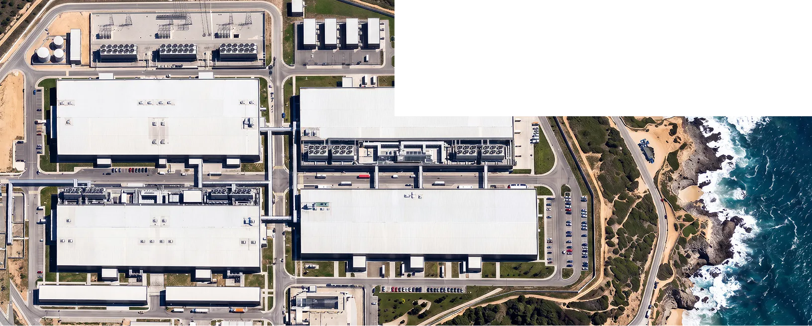 An aerial view of a large industrial complex with several white-roofed buildings, parking lots, and roads beside a rocky coastline with ocean waves.