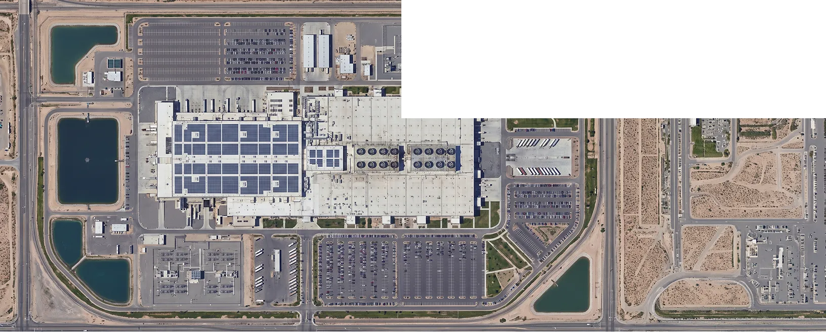 An aerial view of a large industrial facility with rows of solar panels, parking lots, and adjacent water basins beside roads and construction areas.