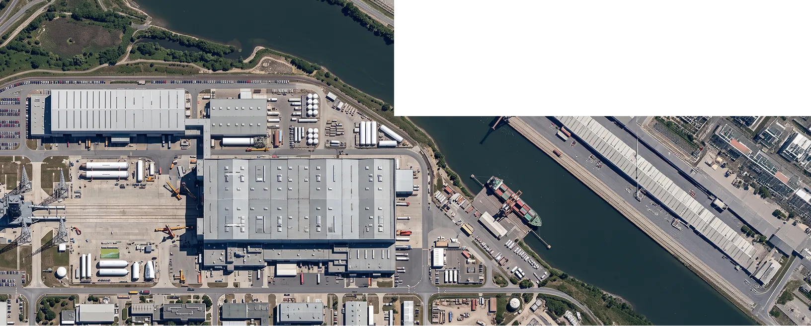An aerial view of a large industrial warehouse complex beside a canal, with docks, trucks, and adjacent buildings.