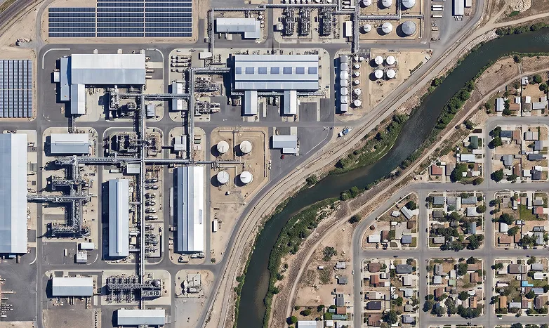 Aerial view of an industrial facility with tanks, buildings, and solar panels beside a river, next to a residential neighborhood.