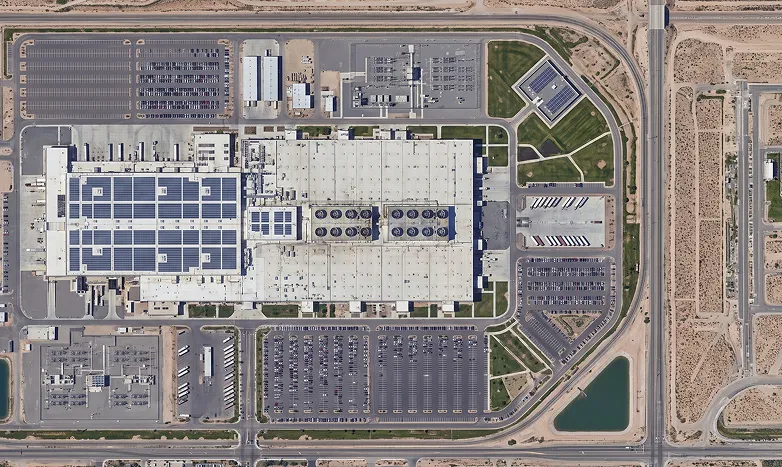 An aerial view of a large industrial complex with rooftop solar panels, parking lots, roads, and surrounding desert terrain.