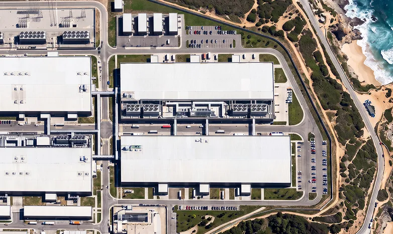 Aerial view of a large industrial complex with white-roofed warehouses, parking lots, and surrounding roads beside a rocky coastline.