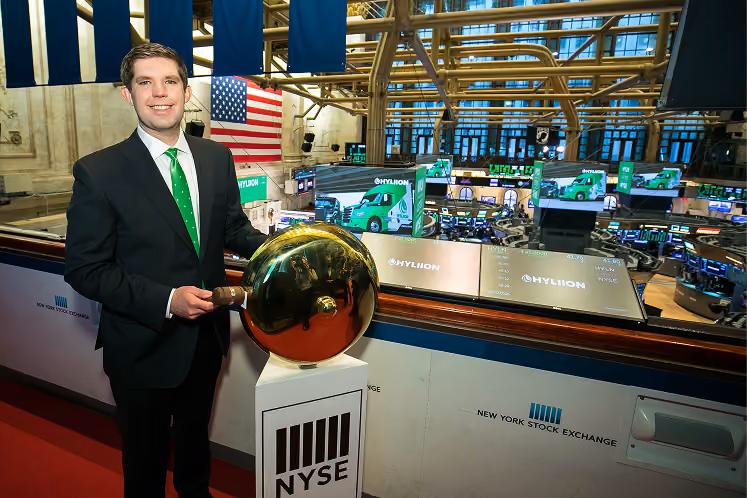 Man in suit rings bell at New York Stock Exchange with trading floor in background.