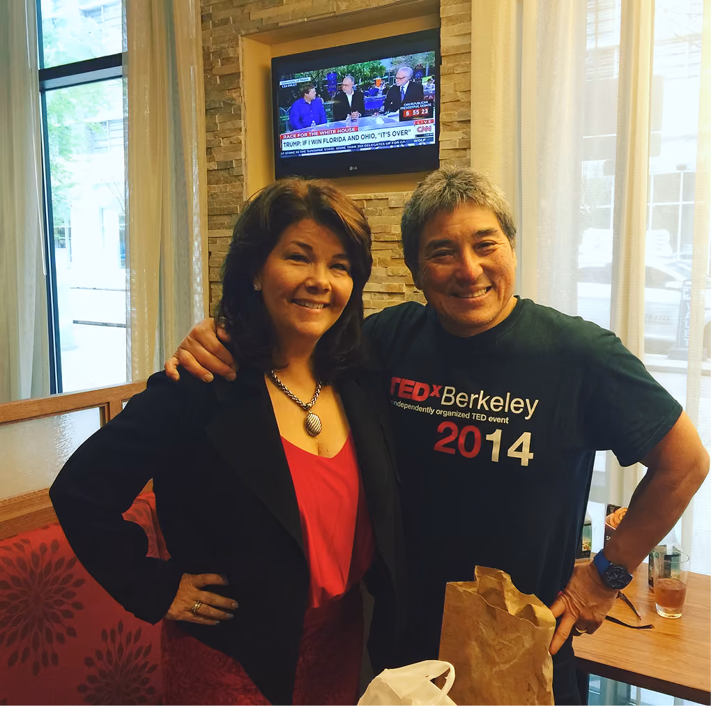 Smiling man in TEDx Berkeley 2014 shirt with arm around woman in red dress and black jacket indoors.