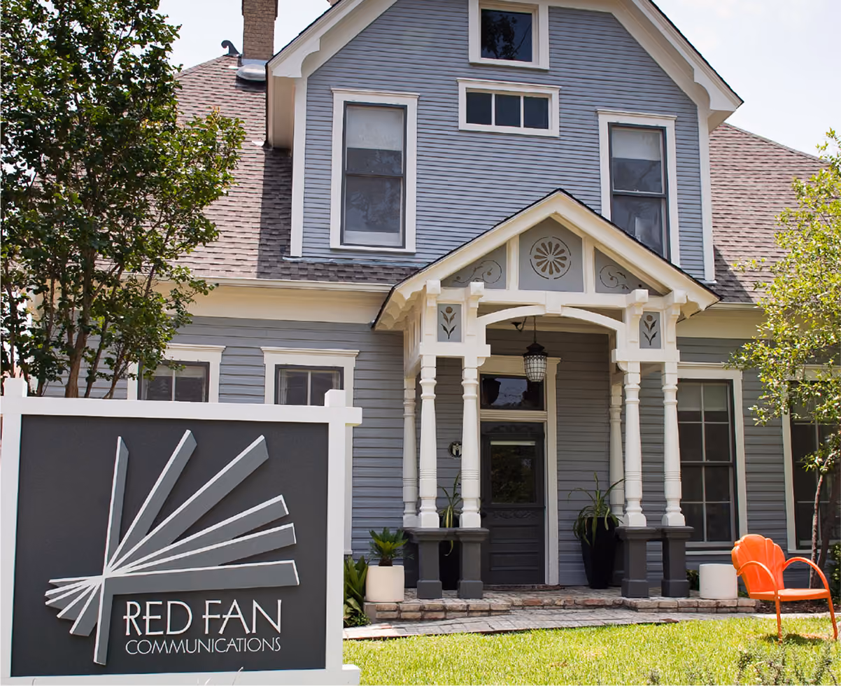 Gray building with white trim and porch, a sign reading Red Fan Communications in front with a red chair on lawn.