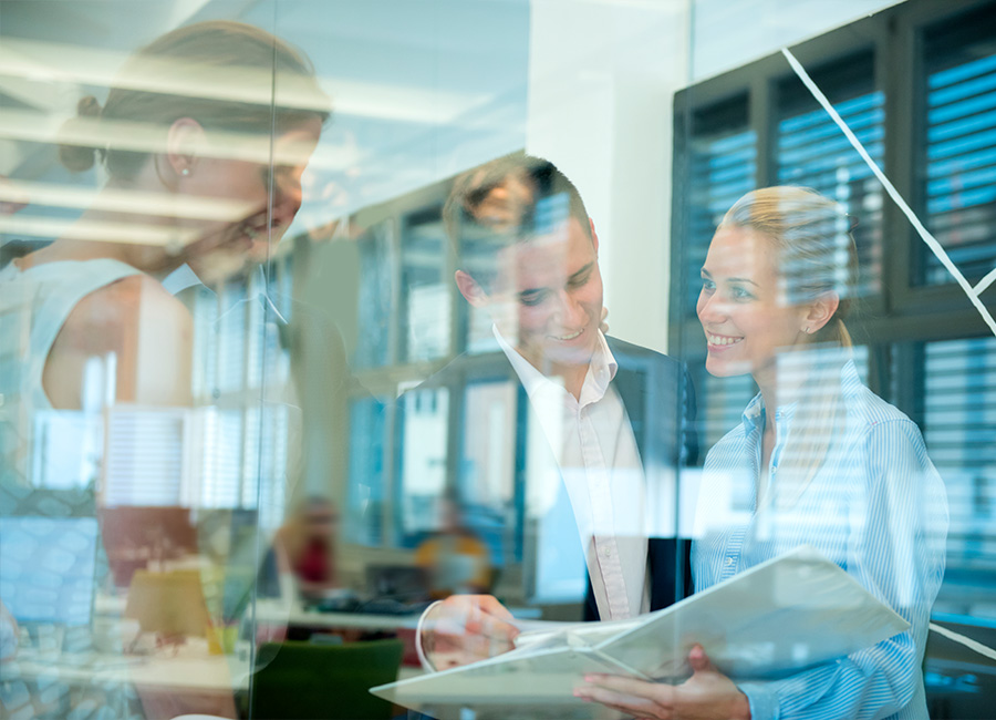 Three business colleagues smiling and reviewing documents together in a modern office setting behind a glass wall.