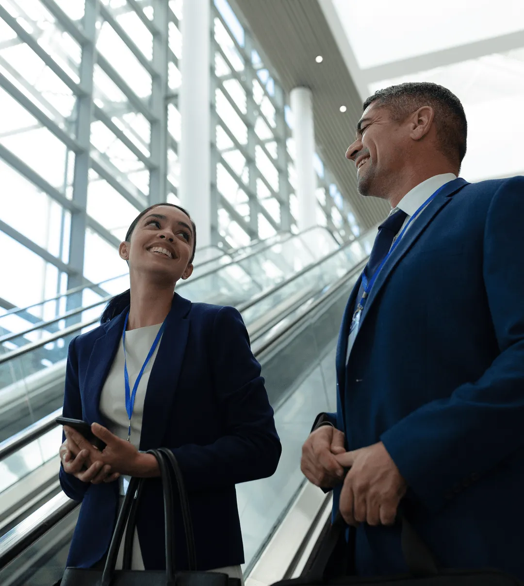 Two business professionals, a woman and a man wearing dark blue suits and conference badges, smiling and talking while standing on an escalator inside a modern glass building.