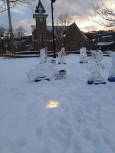 Various ice sculptures with signage noting sponsors on snowy ground, with a clock tower and building in the background
