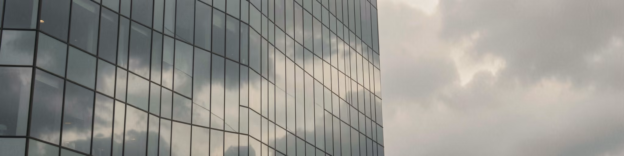 A glass curtain wall of a corporate tower reflecting overcast clouds
