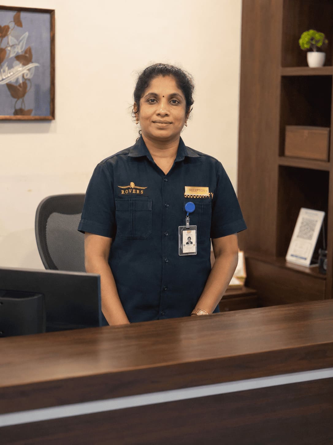 Woman in a dark uniform stands behind a wooden reception desk in an office setting.
