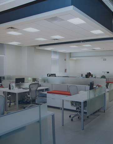 Modern office space with desks, swivel chairs, and frosted glass partitions under fluorescent lights.