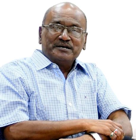 Older man with glasses wearing a blue checkered shirt sitting with arms crossed.