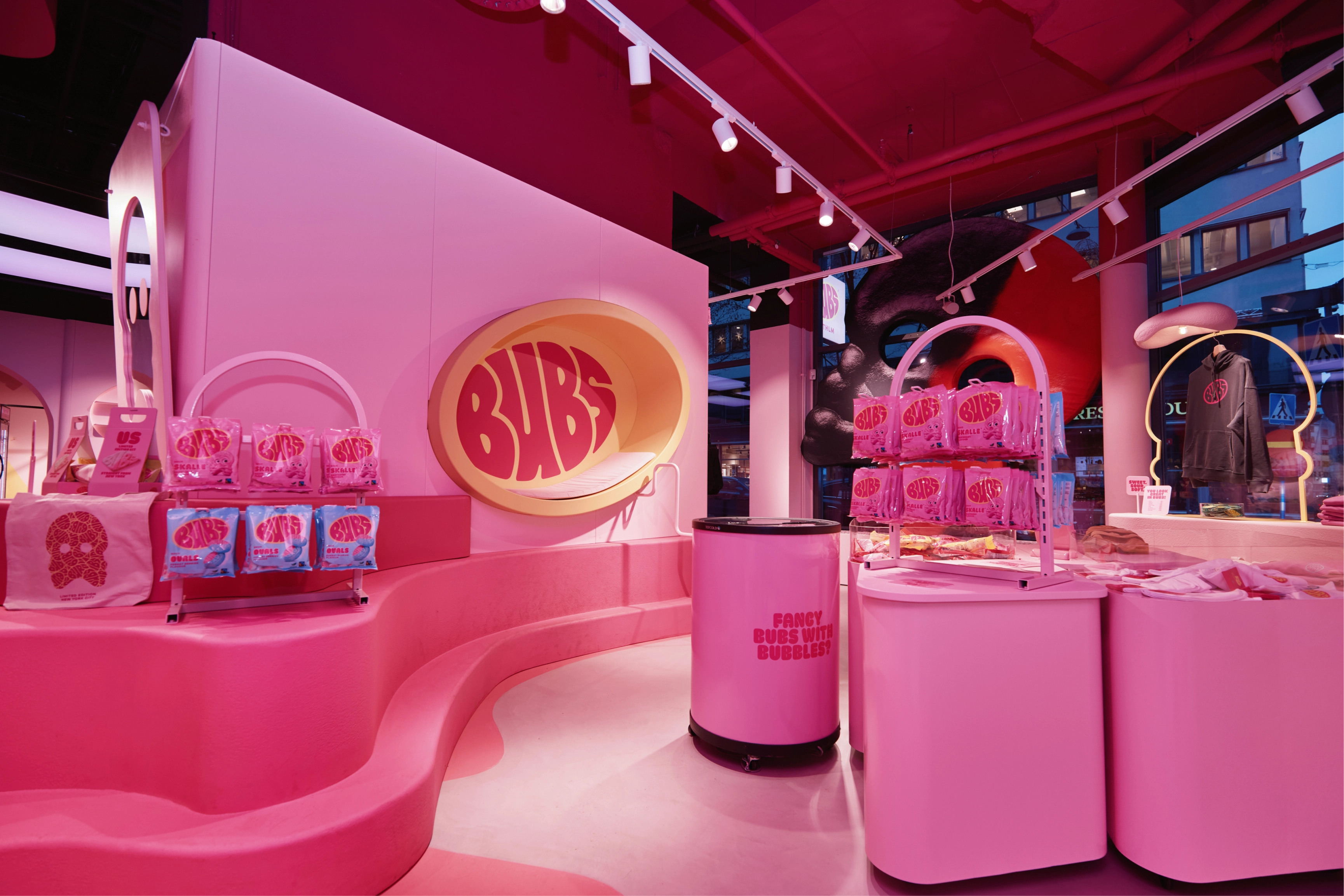 Interior of a BUBS pop-up store in Stockholm, bathed in bright pink lighting. Curved pink display fixtures hold bags of candy, a large glowing BUBS logo sits in a recessed oval on the wall, and a barrel reads "Fancy BUBS with bubbles?" A hoodie hangs in the window on the right.
