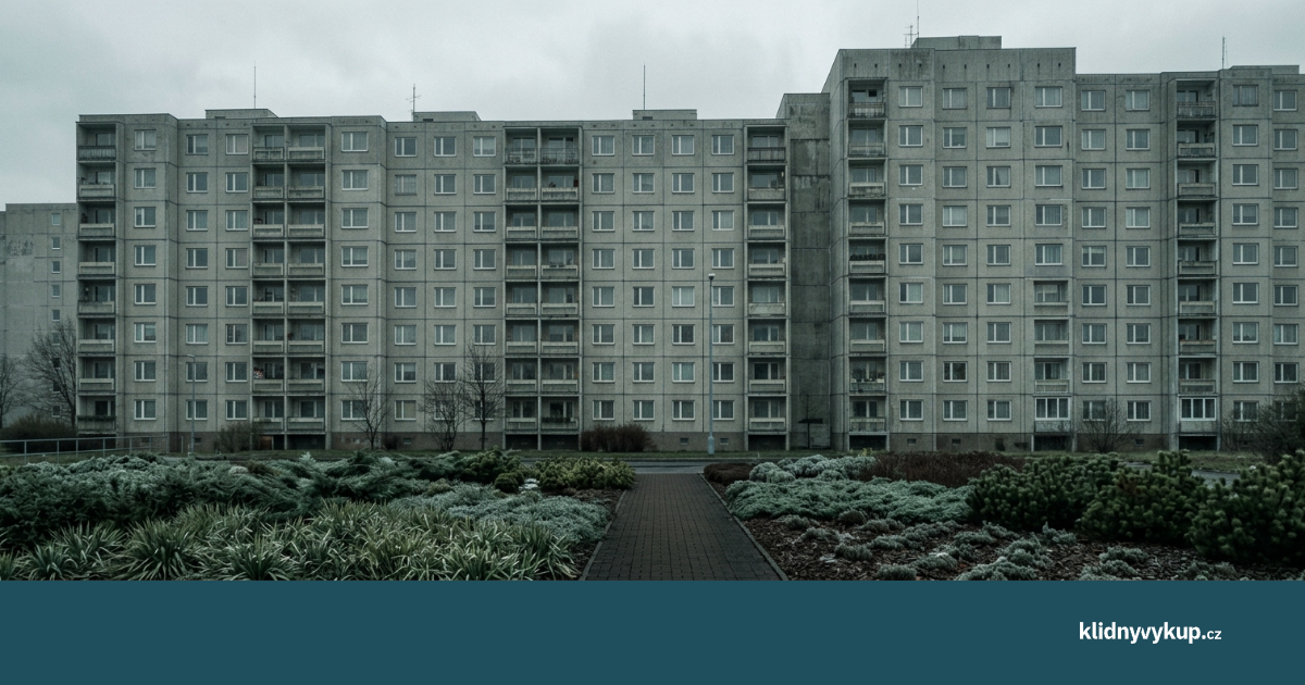 Panelové sídliště přes prázdné parkoviště za zataženého dne.