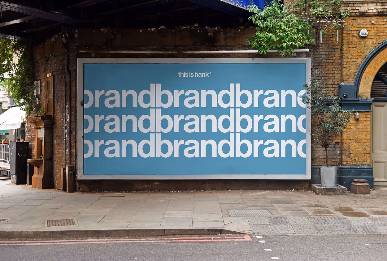 Outdoor billboard on a brick wall under a bridge displaying repeated white text 'brand' in lowercase on a blue background with small text above that reads 'this is hank*'.