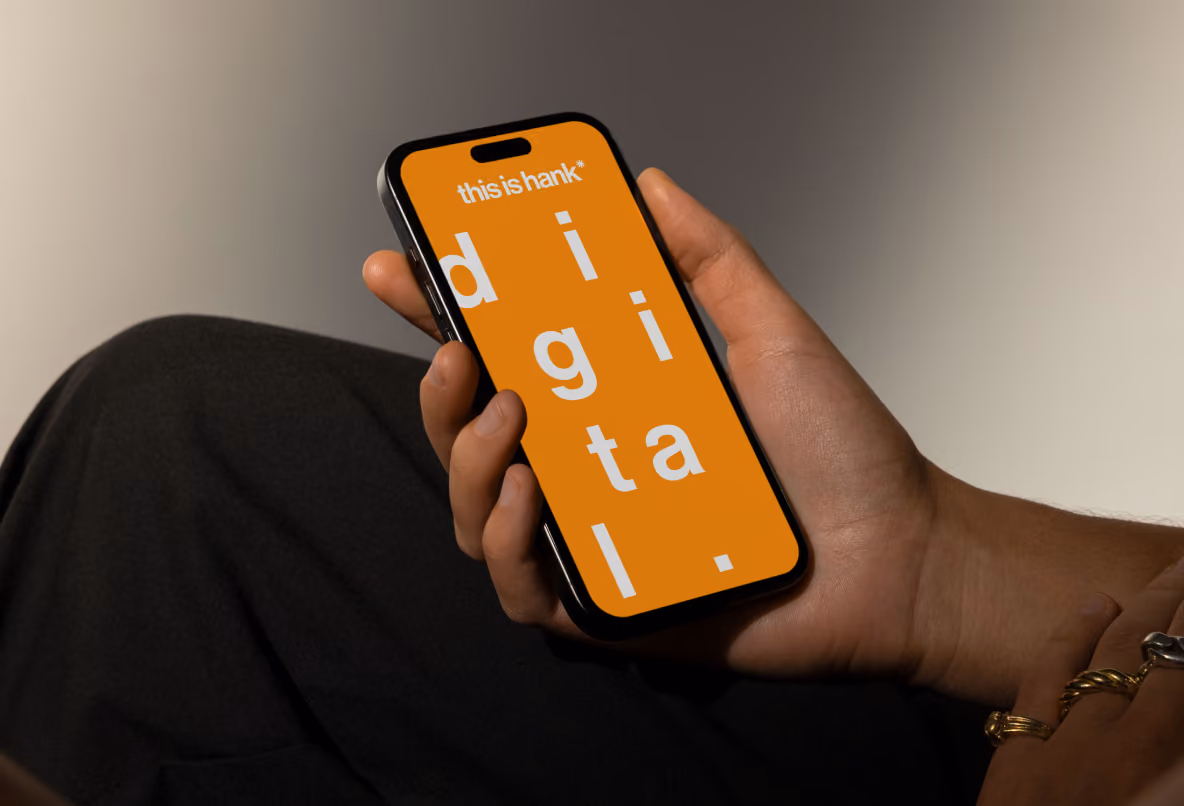 A dark-skinned hand holding a smartphone that displays the word 'digital.' in large white letters on an orange background with 'this is hank' text at the top.