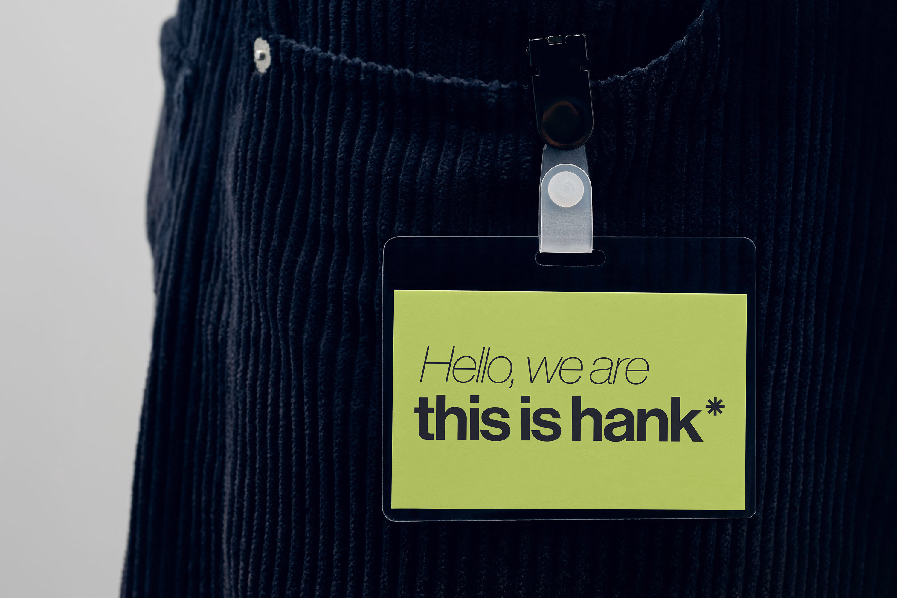 Close-up of a clipped green badge on black corduroy pants reading 'Hello, we are this is hank*' in mixed italics and bold font.