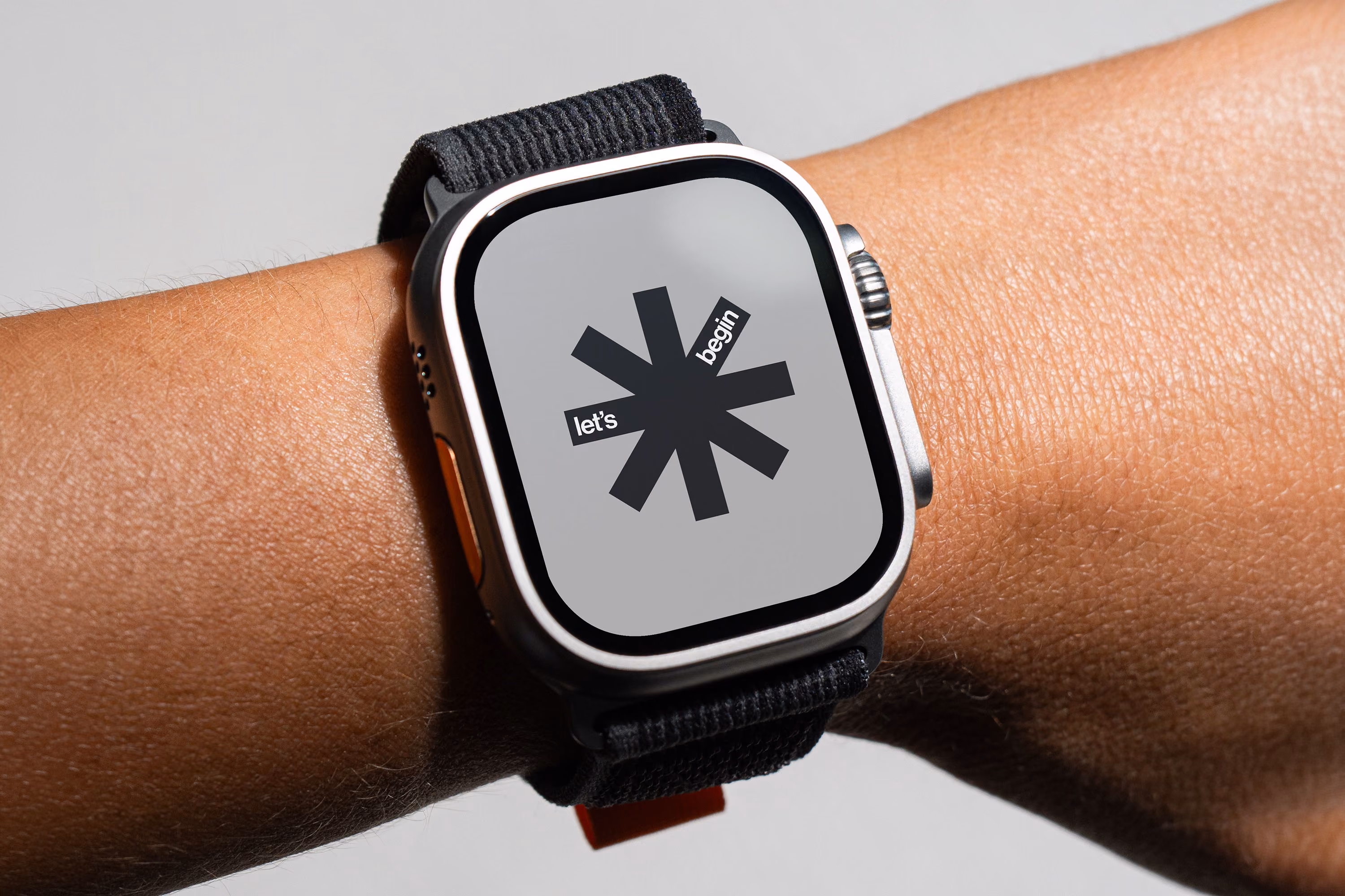 Smartwatch on a wrist displaying a graphic with a black asterisk in the center and the words "let's begin" placed within two of the asterisk's arms on a light gray background.