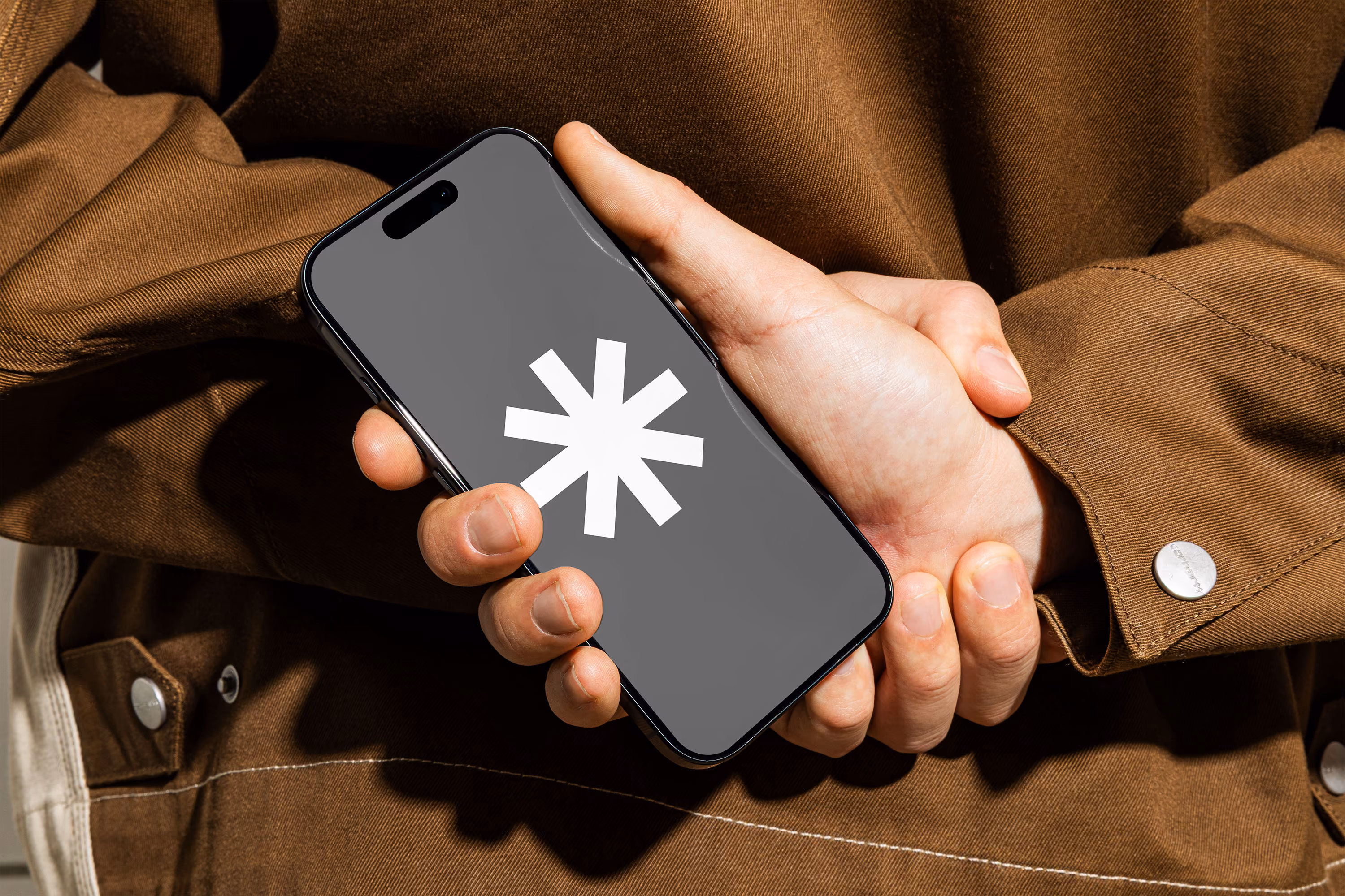 Hand holding a black smartphone displaying a large white asterisk symbol on the screen, against a background of a person wearing a brown jacket.