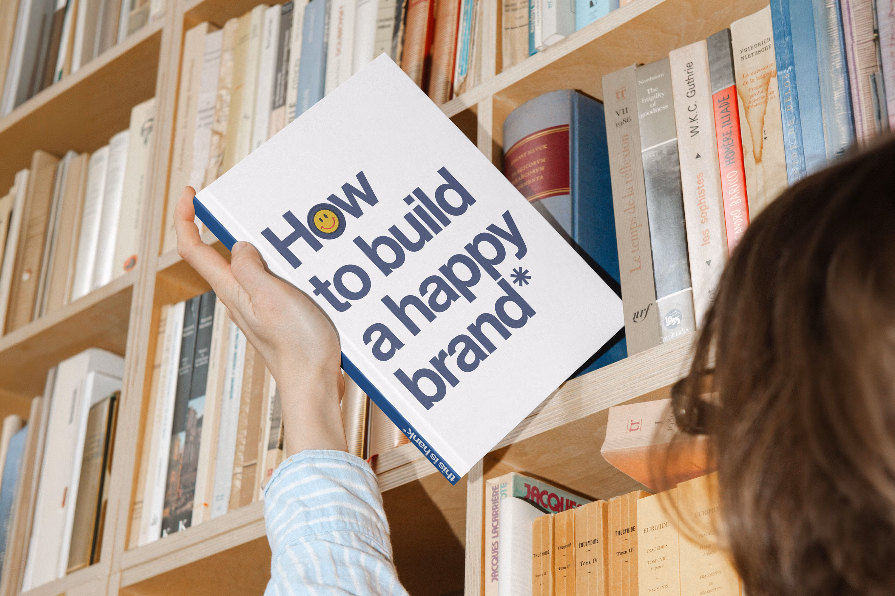 Person's hand holding a book titled 'How to build a happy brand*' in front of a bookshelf filled with various books.