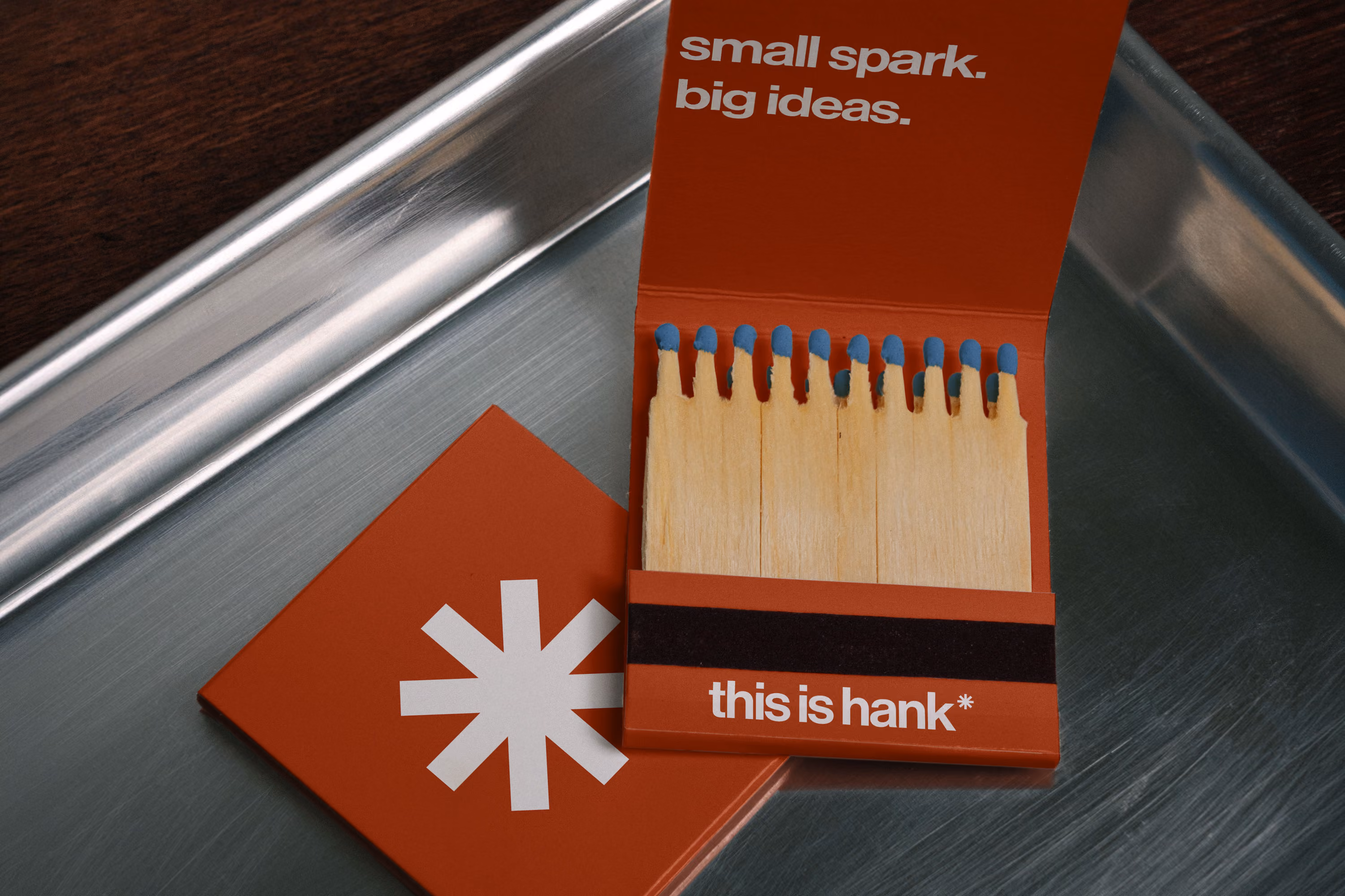 Open matchbook with wooden matches showing blue tips, placed on a metal tray next to closed matchbook with a white asterisk design, inside cover text reads 'small spark. big ideas.' and base reads 'this is hank*'.