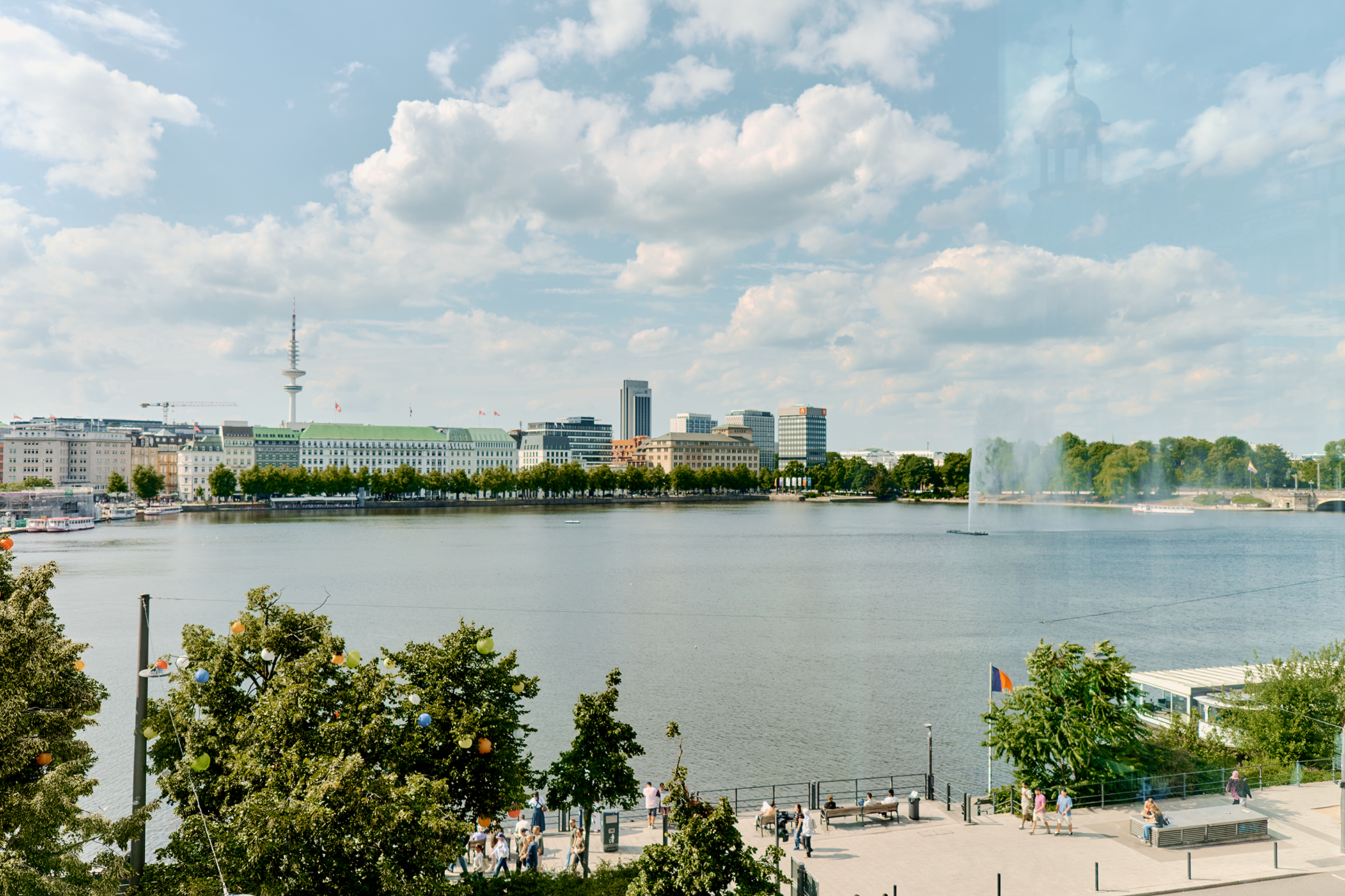 Blick über die Alster mit Springbrunnen, Bäumen und Menschen an der Uferpromenade in Hamburg.