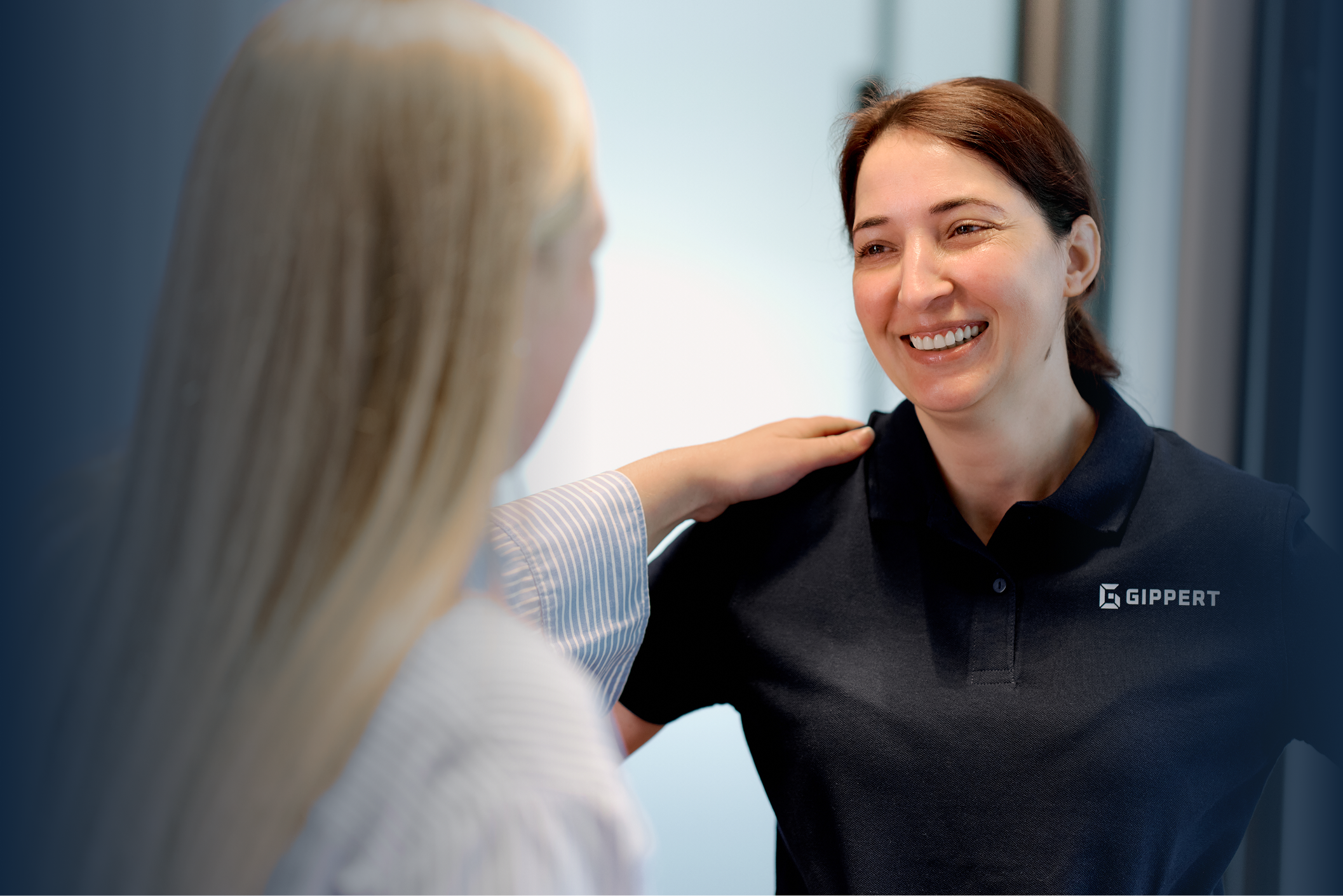 Zwei Frauen im Gespräch, eine legt freundschaftlich Hand auf die Schulter der anderen mit GIPPERT-Logo auf Shirt.