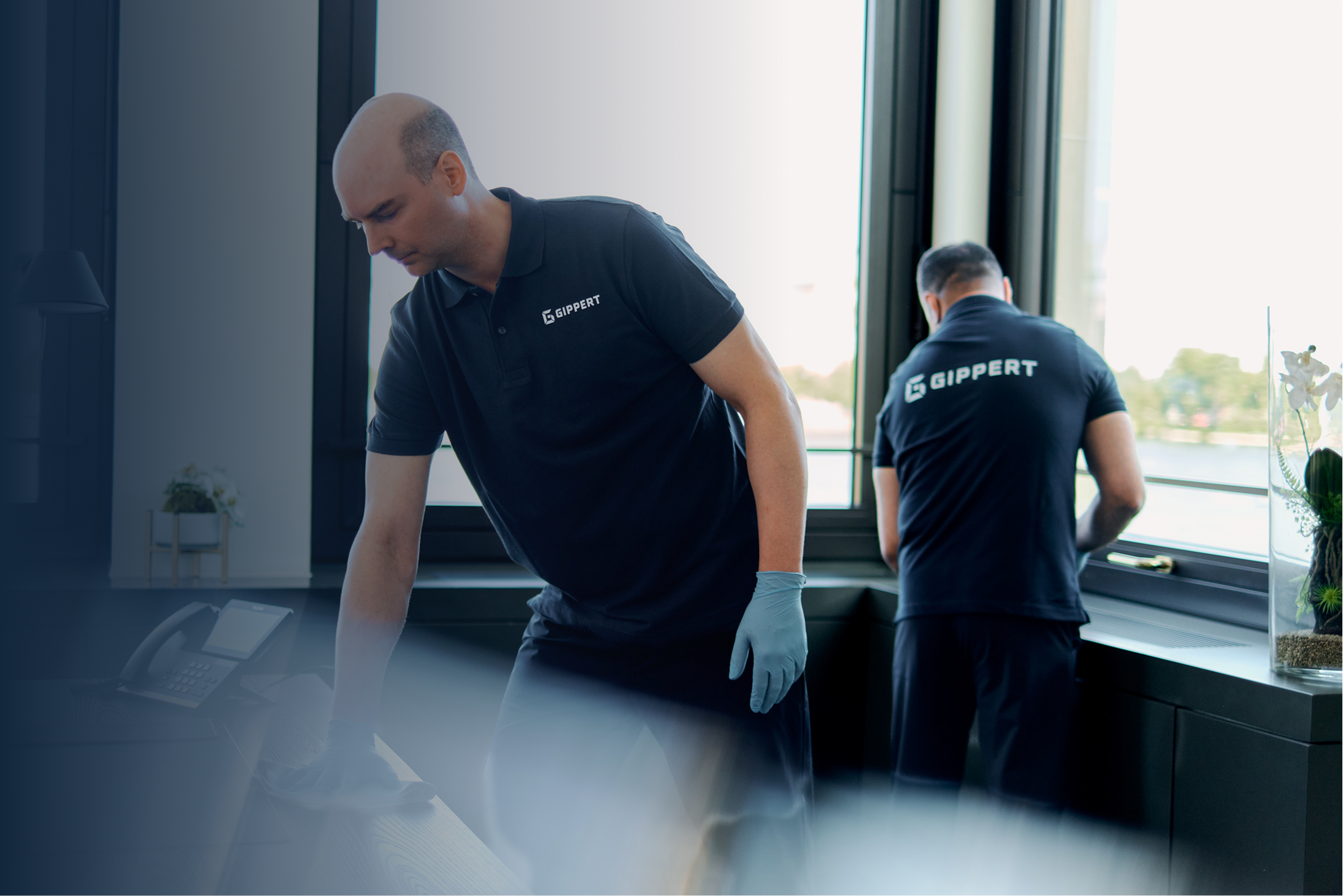 Zwei Männer in schwarzen Gippert-Shirts reinigen Oberflächen in einem hellen Büro mit Fensterblick.
