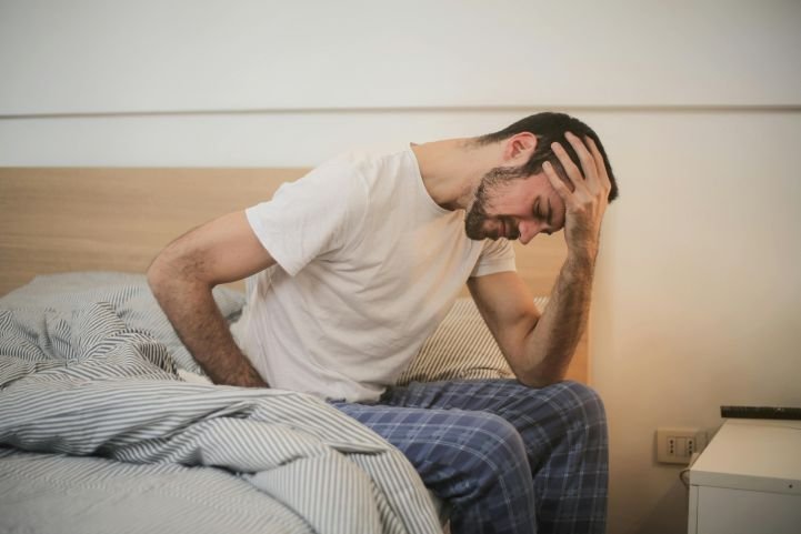 A man sitting on the edge of a bed, holding his head, feeling a panic attack hangover.