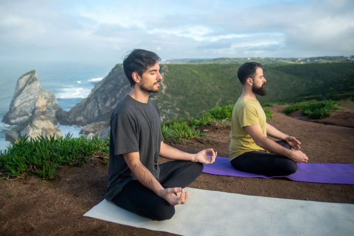 Two guys sitting on yoga mats, meditating in nature, overlooking cliffs and the ocean.