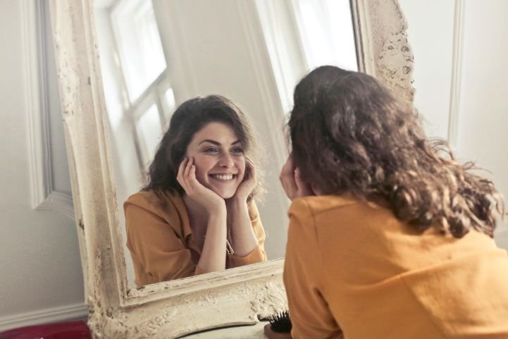 A young woman smiling at her reflection in a mirror, feeling happiness and life satisfaction.