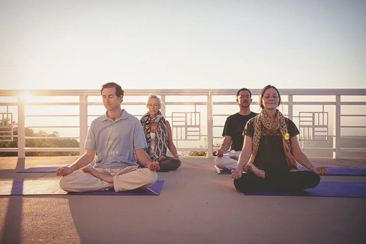 A small sangha is doing a sitting meditation outside on a deck overlooking a sunrise.