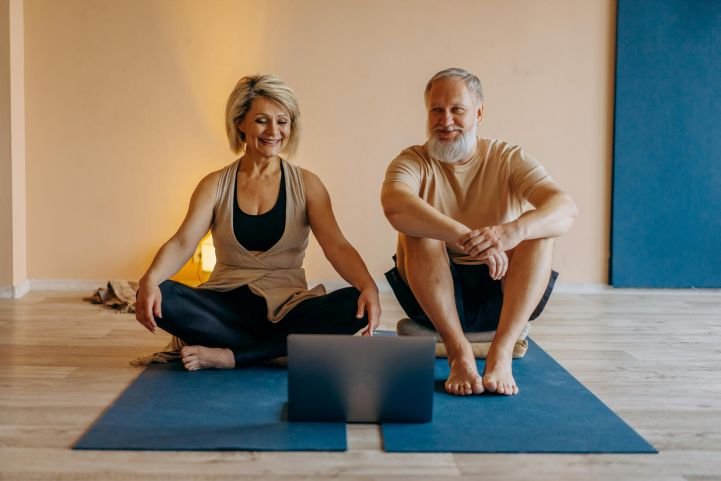 An older couple sitting on yoga mats, enjoying a digital sangha on a laptop.