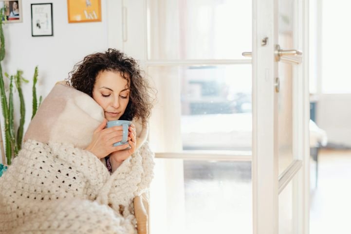 A woman in a cozy blanket enjoying a cup of cocoa near an open door.