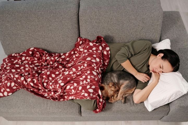 A young woman napping on a couch with a small dog and a red and white blanket.
