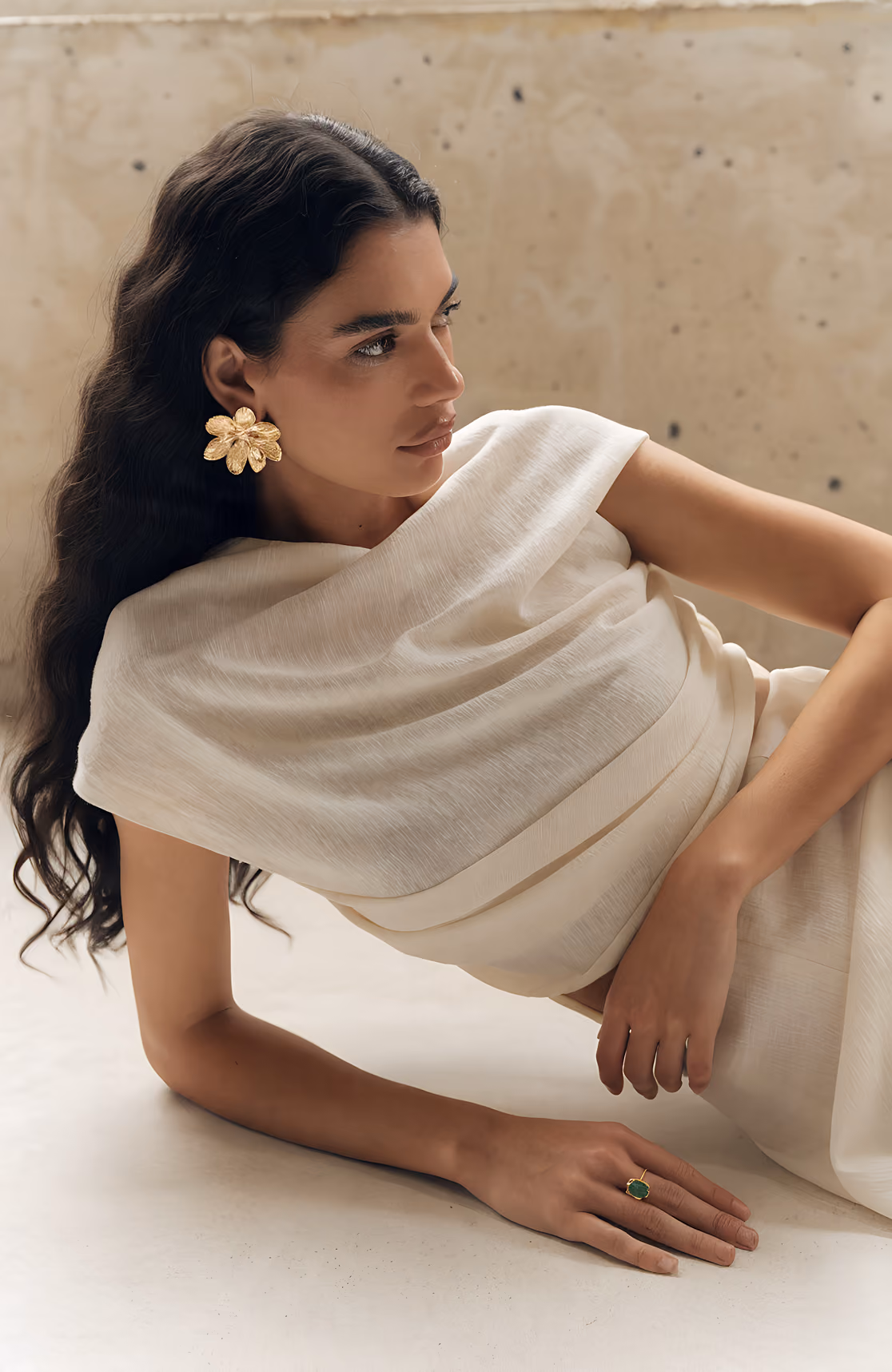 Woman with long dark hair reclines, wearing a white dress, gold flower earring, and green stone ring.