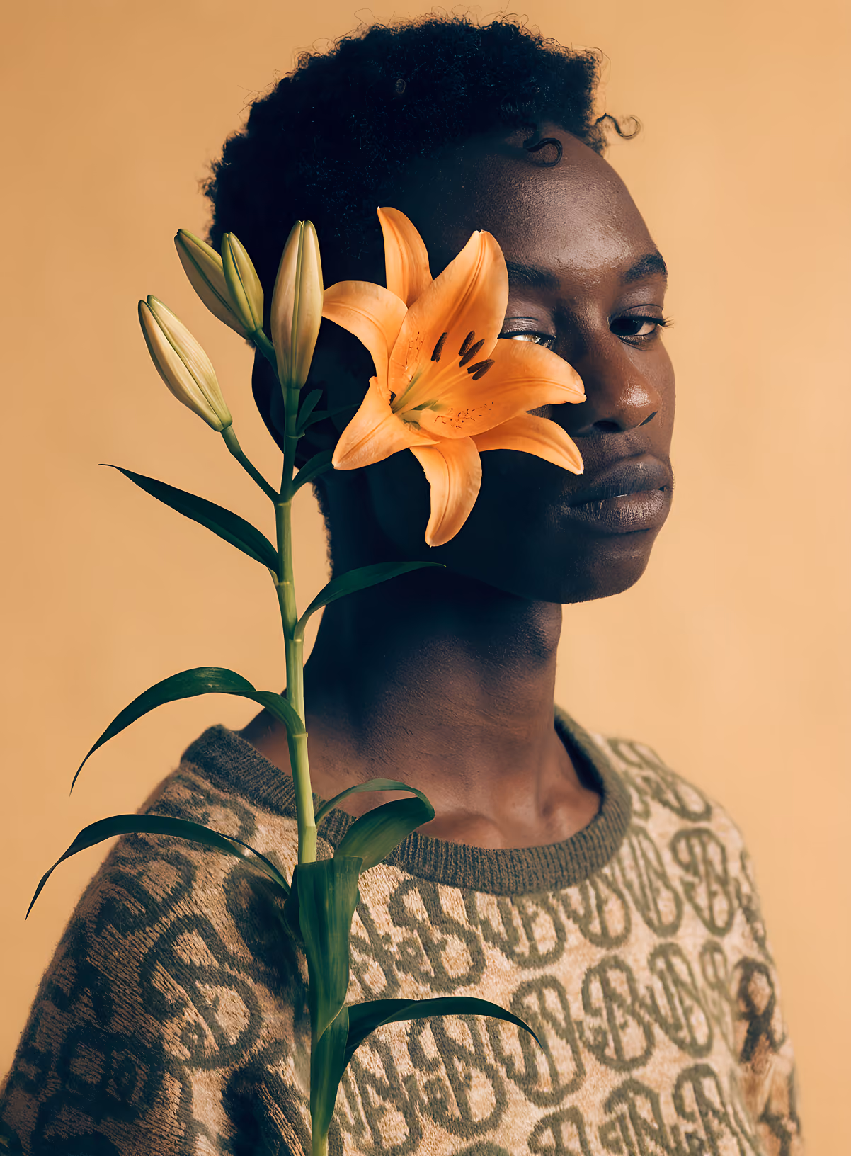 Young person with short curly hair wearing patterned sweater, holding an orange lily flower near face.