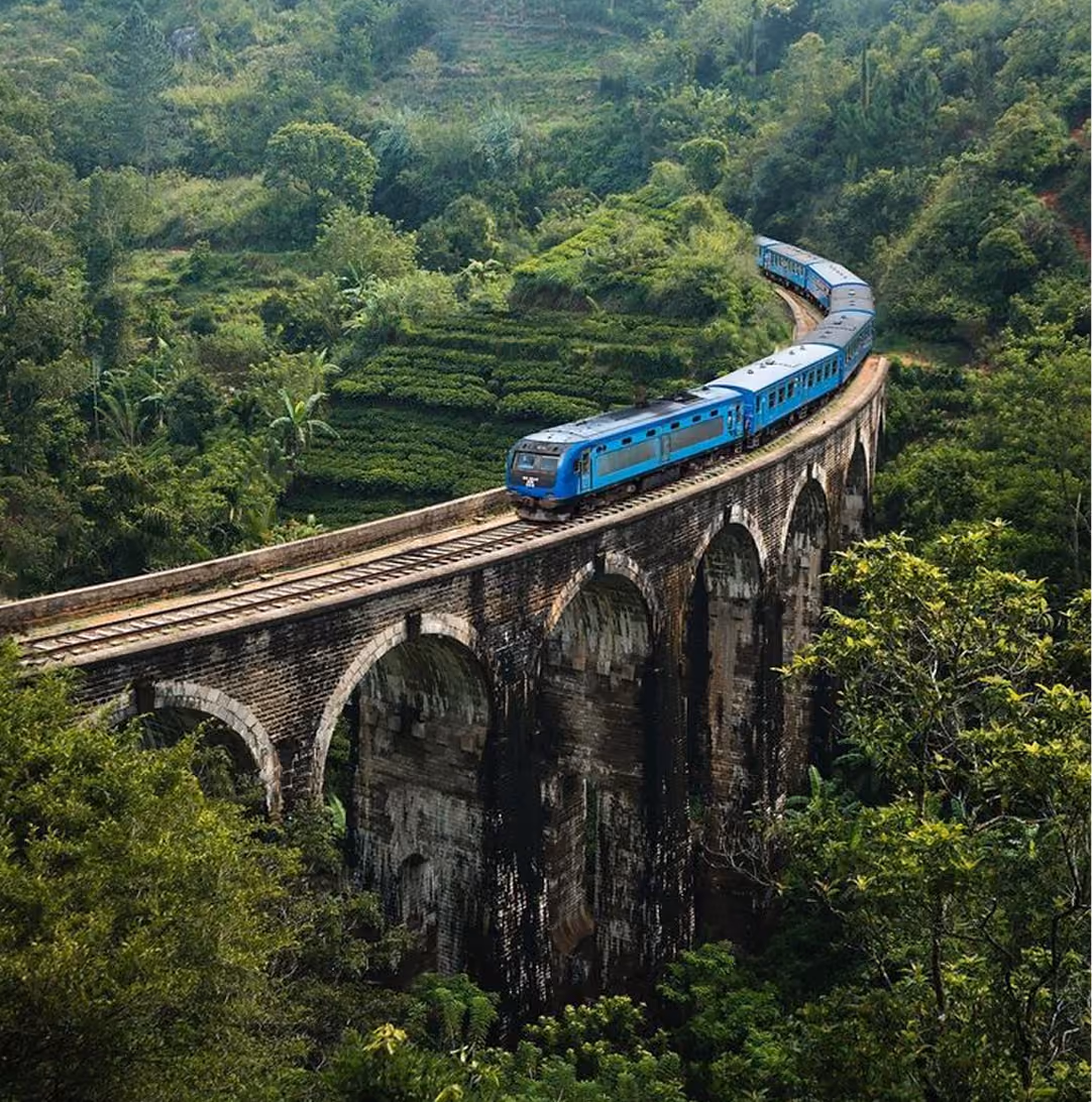 Blue train crossing a tall, arched stone bridge surrounded by dense green foliage and forested hills.