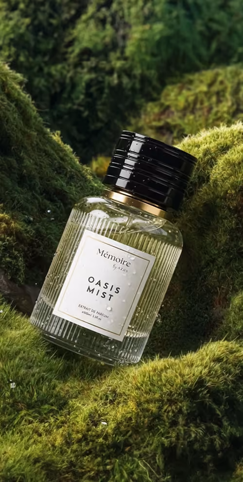 A clear ribbed glass perfume bottle labeled Mémoire Oasis Mist with a black cap, placed on green moss outdoors.