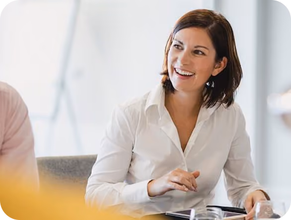 Smiling woman in a white shirt sitting at a table, engaged in conversation in a bright office setting.