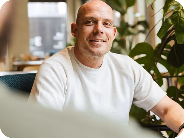 Smiling bald man wearing a white shirt sitting indoors near green plants with a blurred window and furniture in the background.