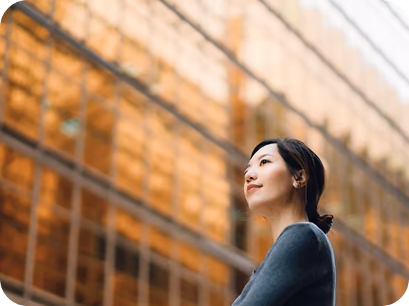 Young woman with dark hair tied back standing outdoors, gazing thoughtfully against a background of reflective orange-tinted glass building panels.