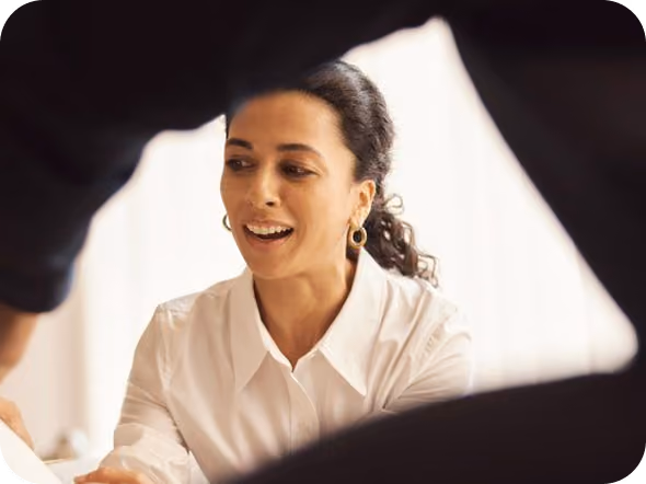 Smiling woman with dark curly hair wearing a white button-up shirt and hoop earrings, engaged in conversation with two people partially visible in the foreground.
