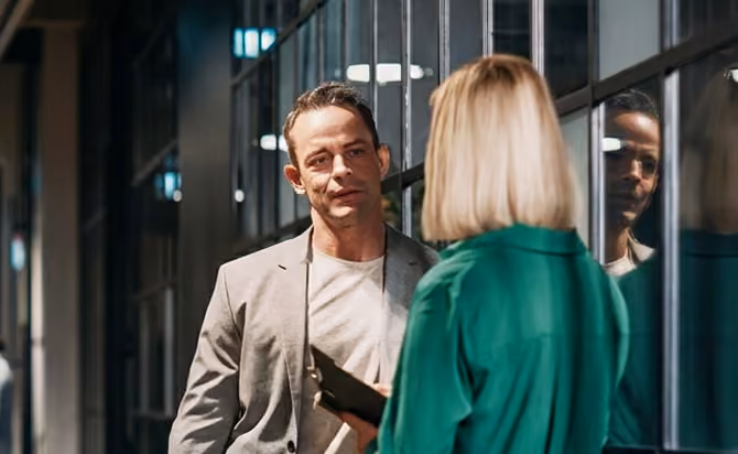 A man in a light gray blazer and light shirt looks at a woman with blonde hair wearing a green jacket, who holds a tablet, standing in a dimly lit hallway with glass panels.