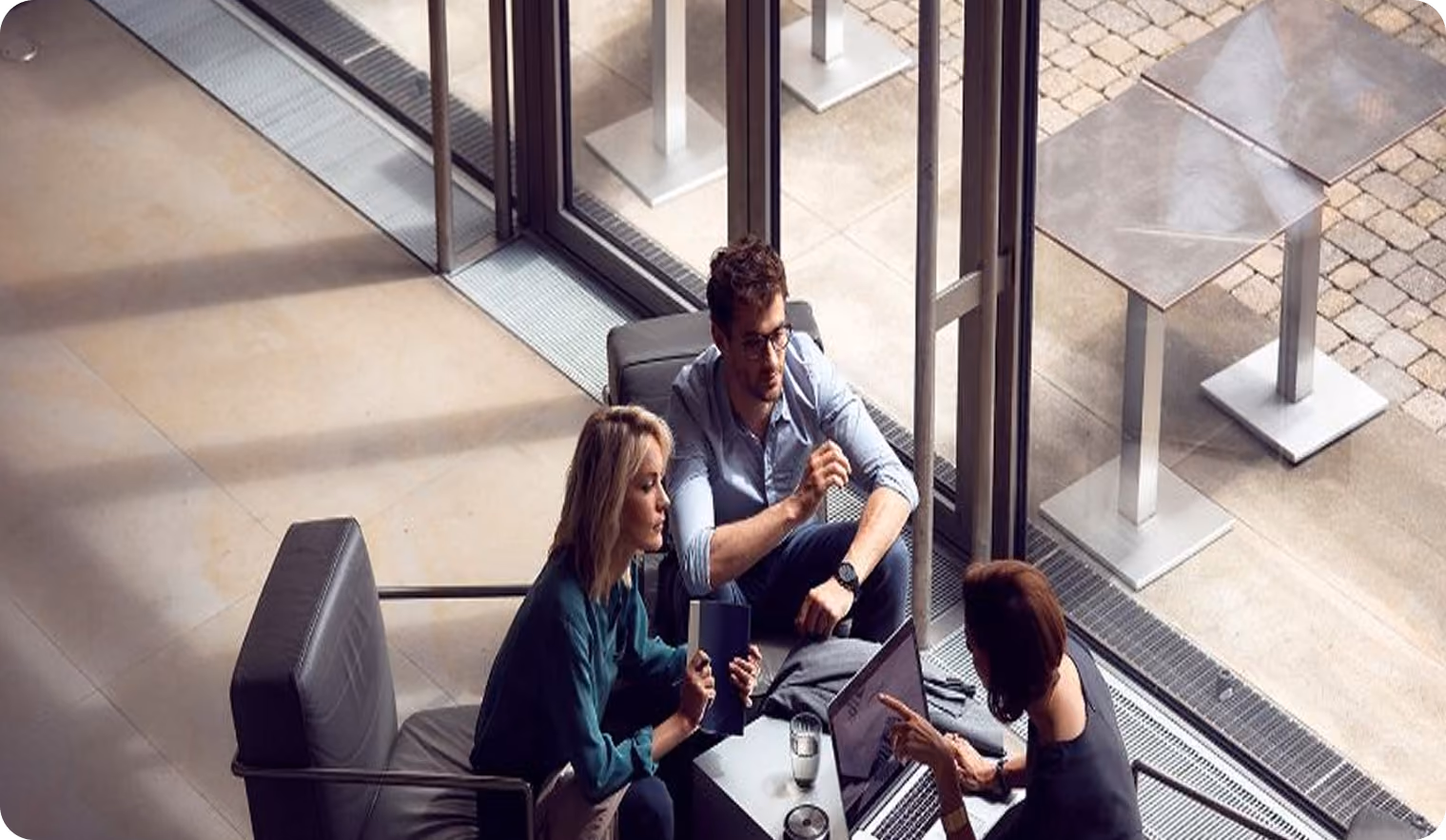 Three people sitting in modern chairs around a glass table in a bright indoor space with large windows, engaged in discussion with a laptop and glass of water on the table.