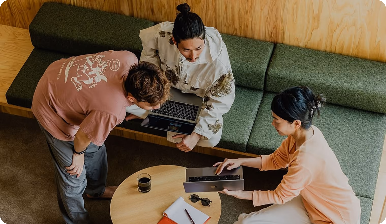 Three people collaborating around a round wooden table with laptops, coffee, a notebook, and sunglasses in a cozy room with green cushioned seating and wooden walls.