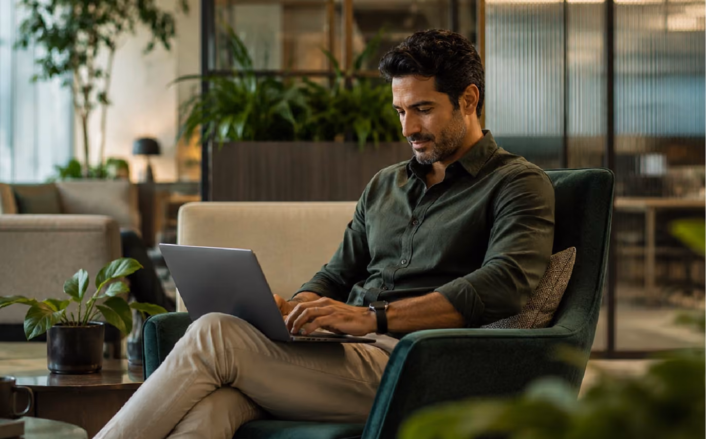 Man sitting in a green armchair typing on a laptop in a modern office with plants and glass partitions.