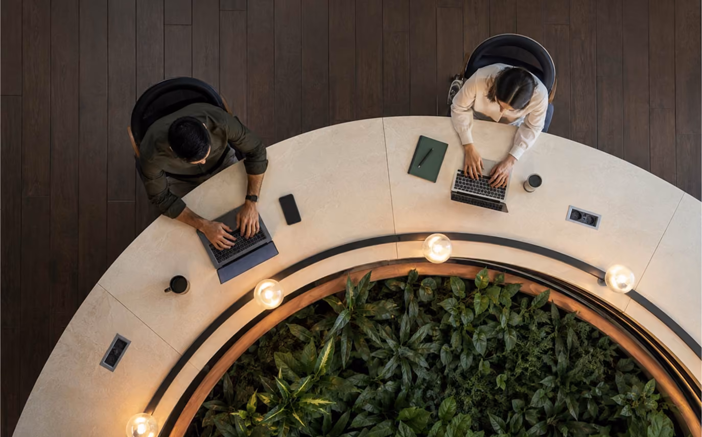 Overhead view of two people seated at a circular table with plants in the center, working on laptops in a modern indoor workspace.