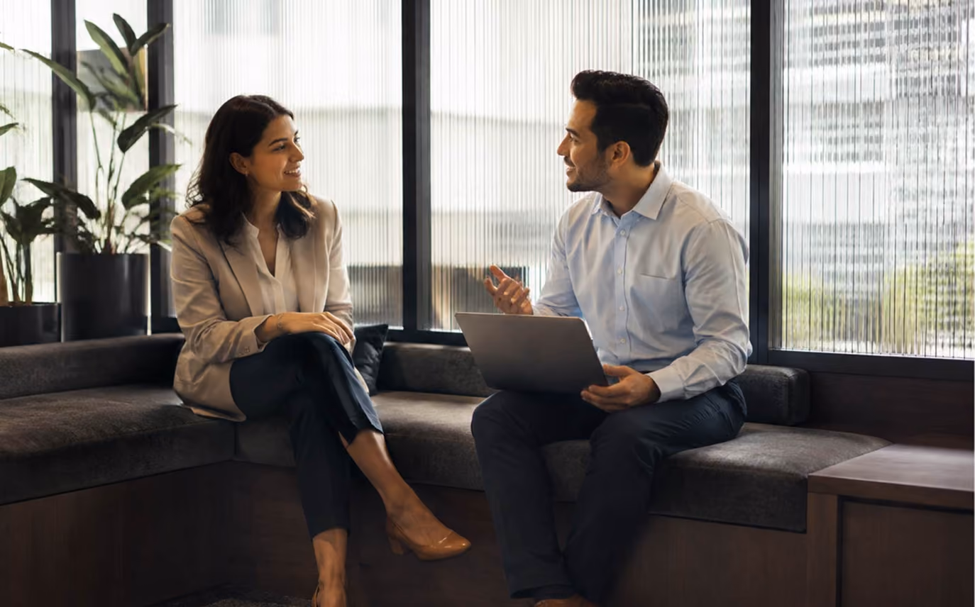 A man and a woman sitting on a cushioned bench in an office space, engaged in a conversation, with the man holding a laptop and gesturing with his hand.