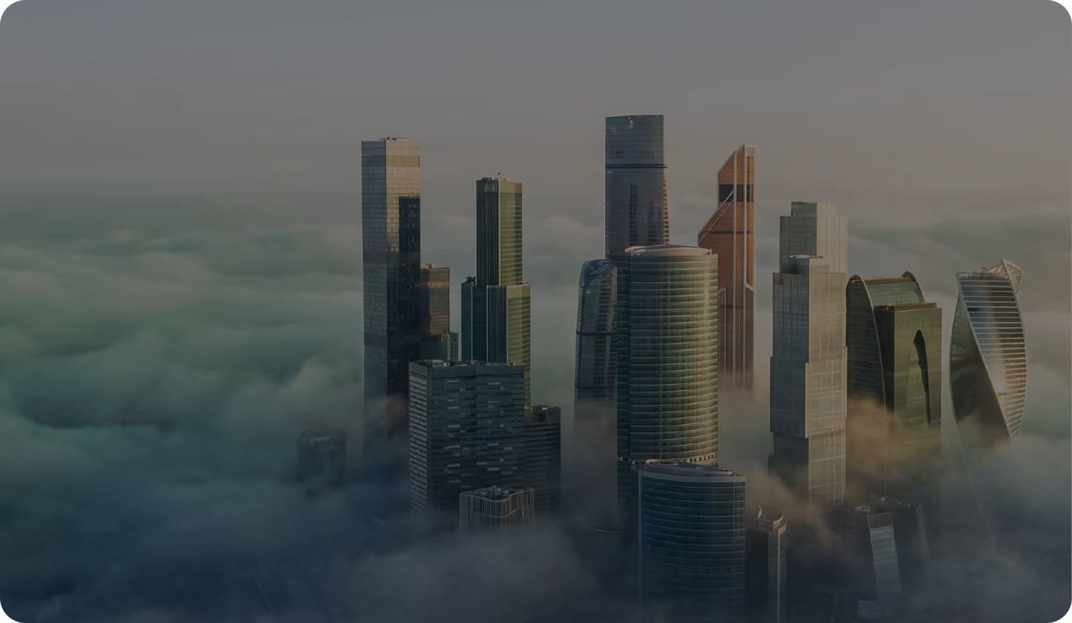 A group of modern skyscrapers partially emerging from thick fog or low clouds during sunrise or sunset, with soft light casting shadows and highlights.